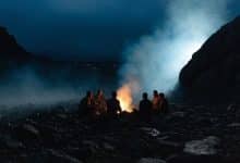 Uma comunidade isolada reunida à noite em torno de uma fogueira, com figuras em silhueta, uma paisagem misteriosa e clima de suspense psicológico.
