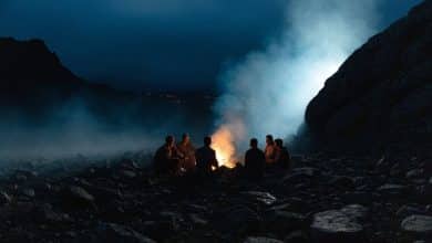 Uma comunidade isolada reunida à noite em torno de uma fogueira, com figuras em silhueta, uma paisagem misteriosa e clima de suspense psicológico.
