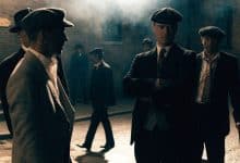 Retrato cinematográfico de um grupo fictício de gângsteres britânicos dos anos 1920 em uma rua industrial de tijolos, com ternos elegantes, boinas, fumaça e iluminação dramática.
